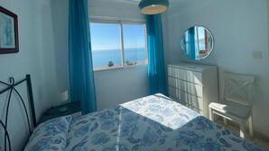 A bedroom with a bed, a window with curtains, a dresser, a mirror, and a view of the sea. - TERRASOL FRONTAL AL MAR ANTILLAS 8 (Torre del Mar)