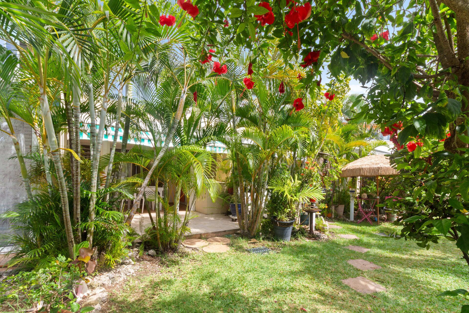 Un exuberante jardín con palmeras y flores rojas, una estructura con techo de paja y un camino pavimentado.