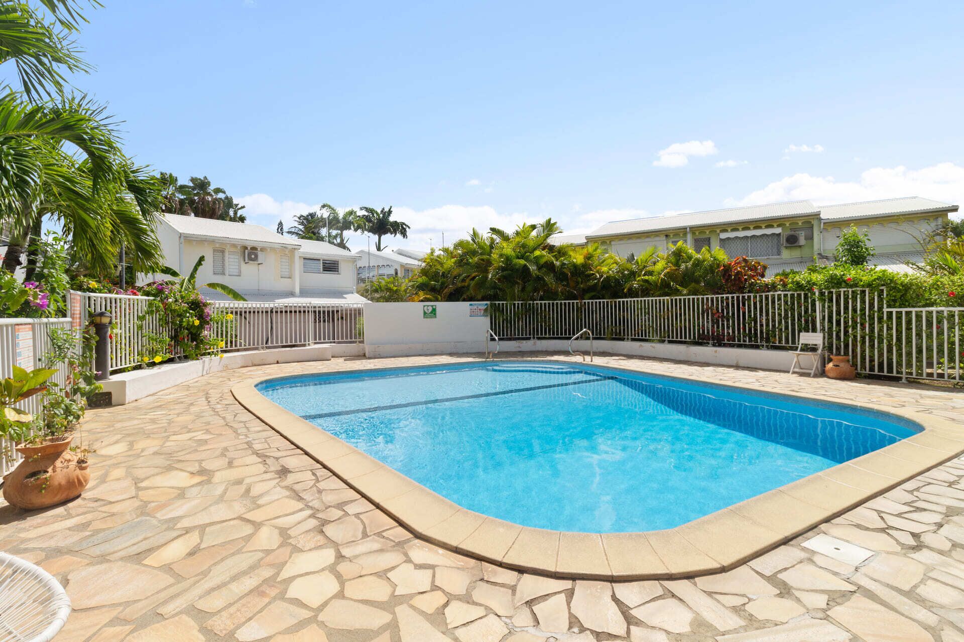 Une piscine rectangulaire bordée de carreaux de pierre, entourée d’une clôture blanche et d’une végétation luxuriante.