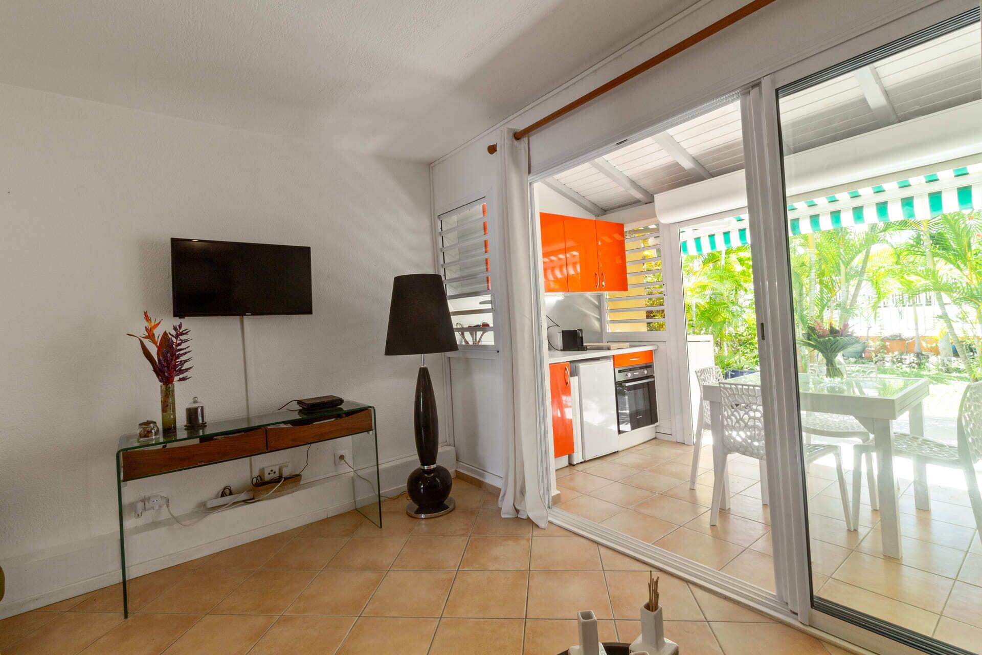 A modern living room with a TV, a glass coffee table, and a sofa. There is a sliding glass door leading to a balcony with outdoor furniture.