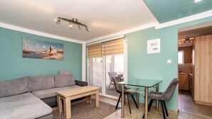 Una sala de estar con un sofá, una mesa de vidrio y sillas. Hay un cuadro en la pared y una puerta que da a una cocina.