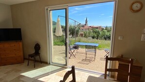 A room with a sliding glass door leading to an outdoor area with a table and chairs, a wooden dresser, a television, and a clock on the wall. - Gite TT Confort Proche Millau / Roquefort VUE Panoramique Piscine Chauffee (SAINT-VICTOR-ET-MELVIEU)