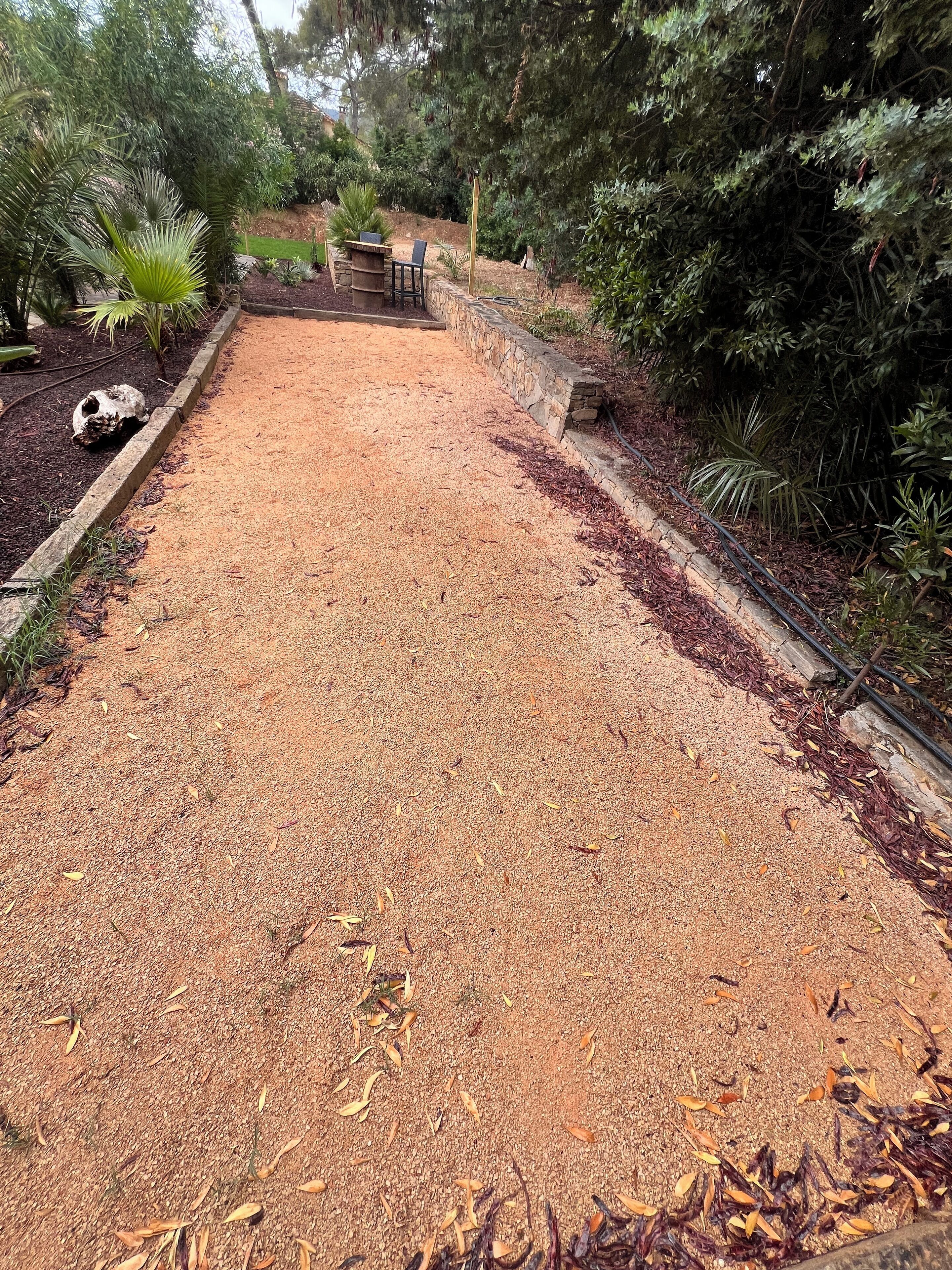 A gravel pathway bordered by stone edging, surrounded by dense foliage and a small seating area with a table and chairs.