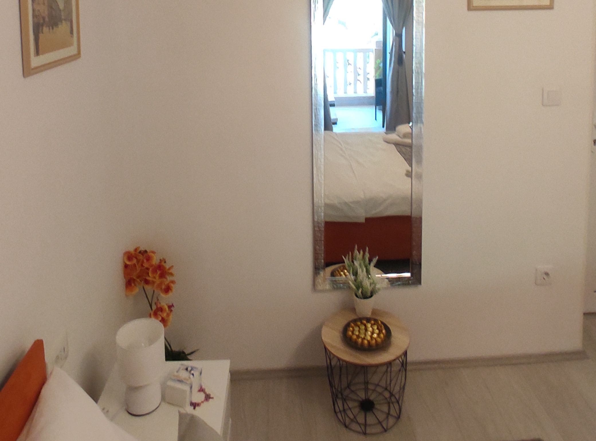 Ein Hotelzimmer mit einem Bett, einem Nachttisch mit Blumenvase, einem kleinen runden Tisch mit einer Schale und einem Spiegel an der Wand.