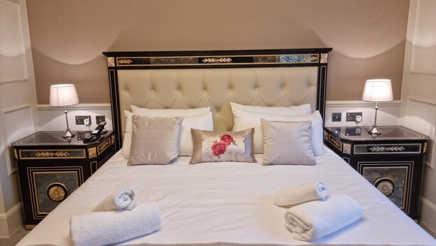 A neatly made bed with white linens and decorative pillows, two bedside tables with lamps, and a classic tufted headboard. - Weston Manor (Bicester)