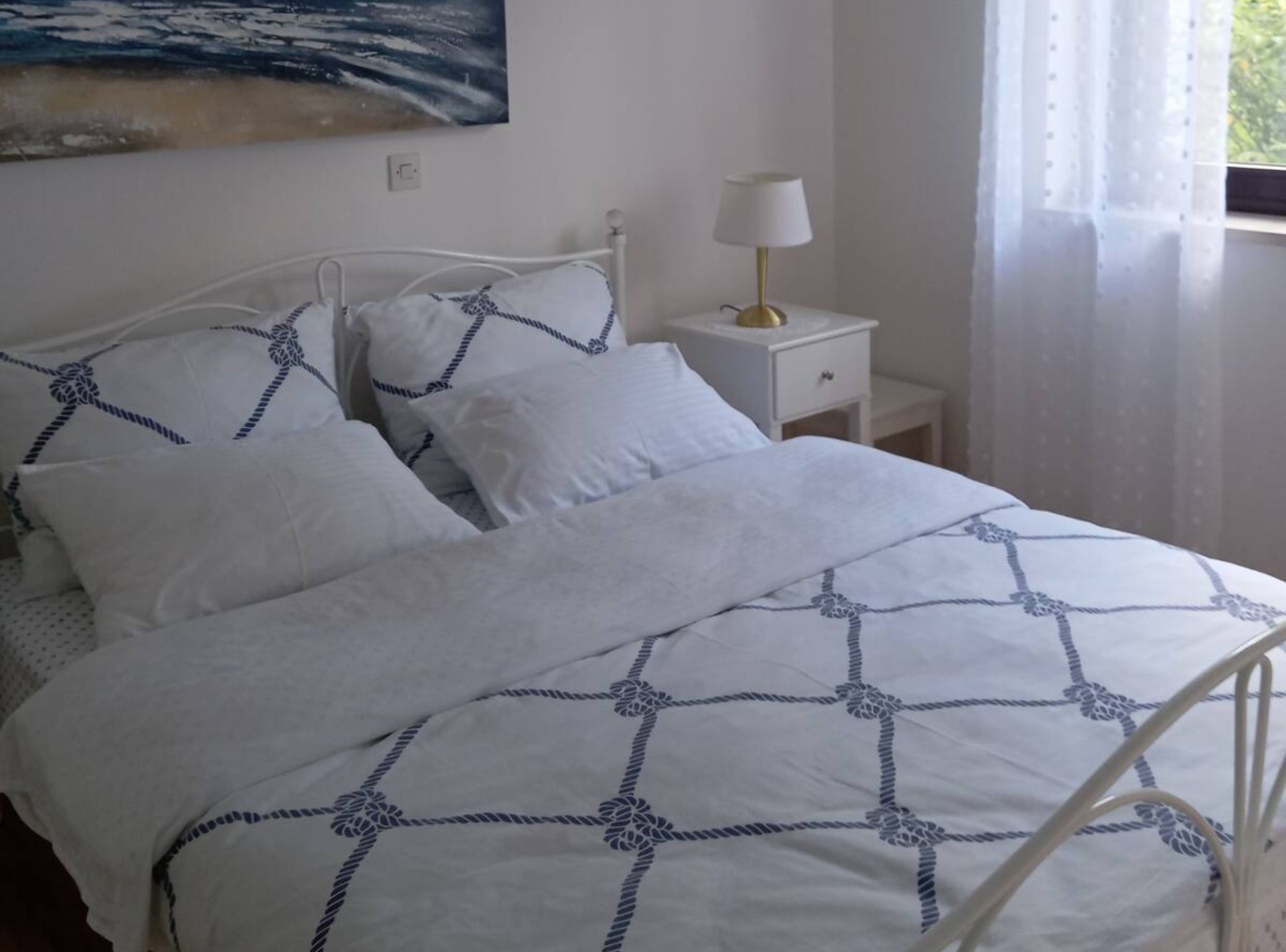 Een net opgemaakt bed met een deken in wit en blauw, een wit metalen hoofdbord en een nachtkastje met een lamp.