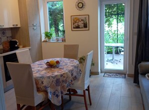 A dining area with a round table covered in a floral tablecloth, surrounded by chairs. There is a bowl of fruit on the table, a clock on the wall, and a window with a view of greenery outside. - Green Garden Oasis (Opatija)