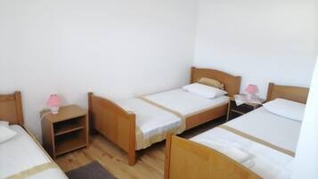 A room with three wooden beds, each with a white mattress and a wooden headboard. There are bedside tables with lamps, and a small shelf unit under one of the beds.