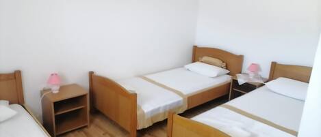 A room with three wooden beds, each with a white mattress and a wooden headboard. There are bedside tables with lamps, and a small shelf unit under one of the beds.
