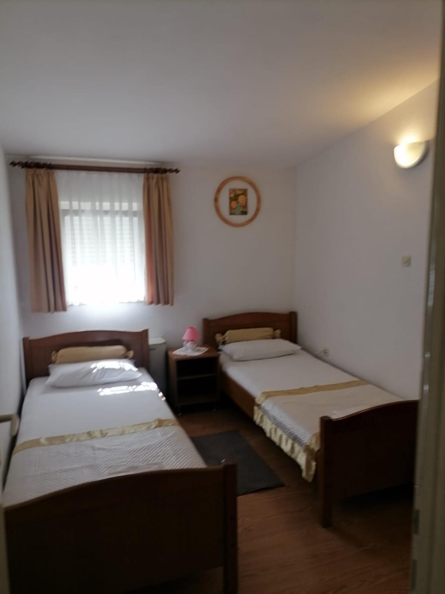 A small, simple room with two single beds, a nightstand, a lamp, and a window with curtains.