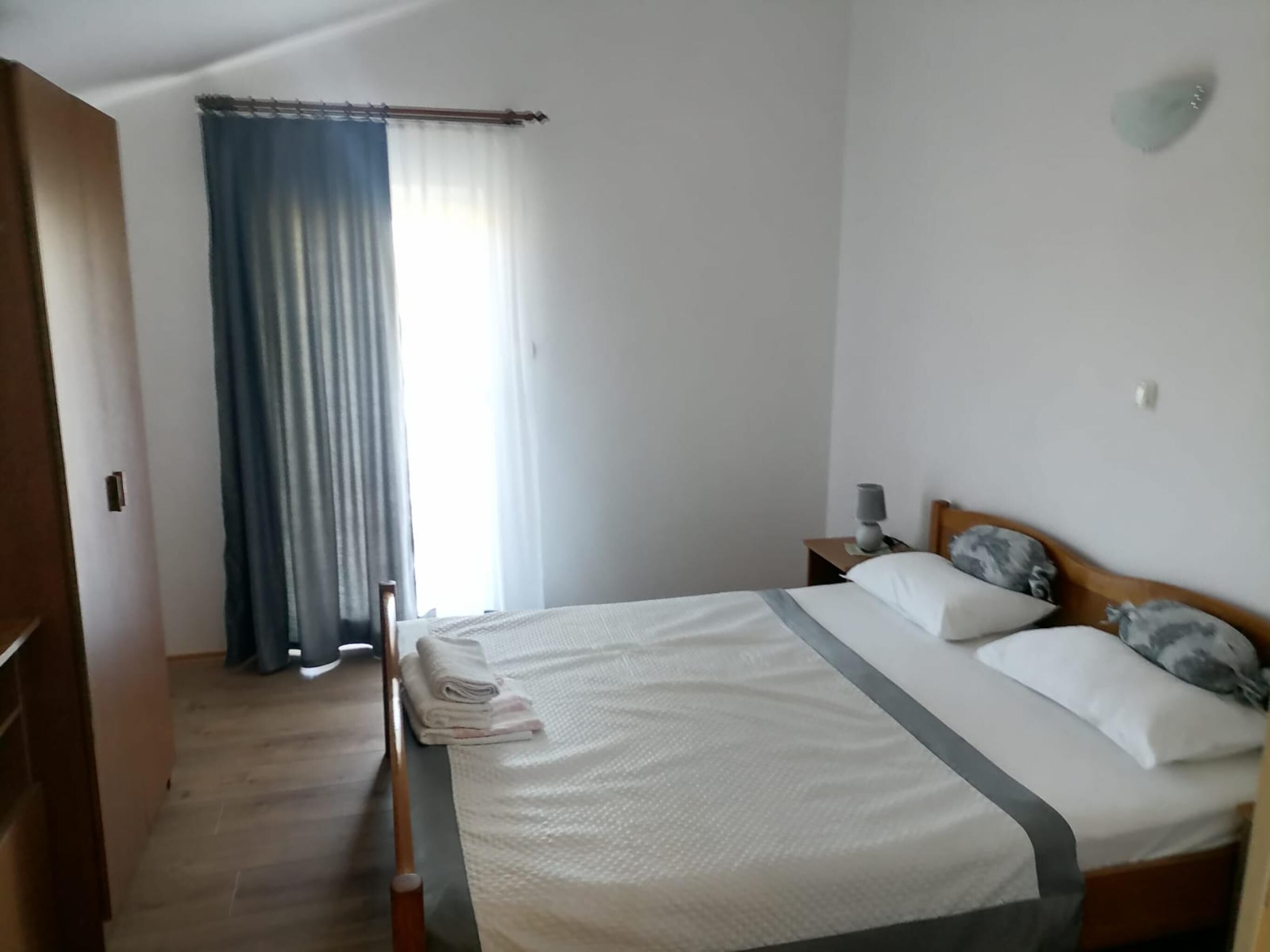 Un dormitorio con una cama, una mesita de noche con lámpara, una ventana con cortinas y una puerta de madera.
