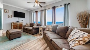 Amplia sala de estar con un sofá grande marrón, un sofá más pequeño y un reposapiés a juego. Hay un televisor de pantalla plana montado en la pared, un mueble de madera y una pequeña mesita. La habitación tiene ventanas amplias con cortinas, un ventilador de techo y vistas al océano.