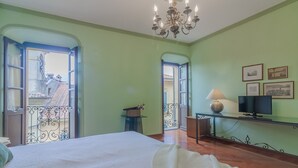 A bedroom with a large bed, a chandelier, a desk with a lamp, and a television. There are open doors leading to a balcony with a view of buildings. - Daisy Home by Varenna Holidays (Varenna)