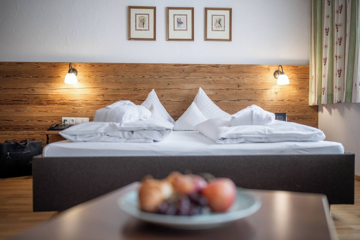 Ein ordentlich bezogenes Bett mit Kissen, ein Nachttisch mit Obstplatte und gerahmte Bilder an der Wand.