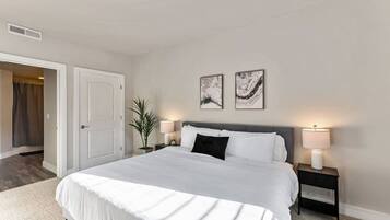 Une chambre à coucher avec un grand lit, deux tables de chevet avec des lampes, une plante en pot et des œuvres d’art abstraites accrochées au mur.