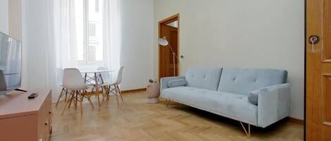 Ein modernes Wohnzimmer mit einem hellblauen Sofa, einem Holzboden im Fischgrätmuster, einer Glastrennwand für den Fernseher und einem Essbereich mit weißen Stühlen.
