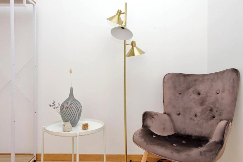 Ein Zimmer mit einem grauen Polsterarmlehnstuhl, einem weißen Beistelltisch mit einer Vase und einer hohen Stehlampe mit goldener Lampenschirm.