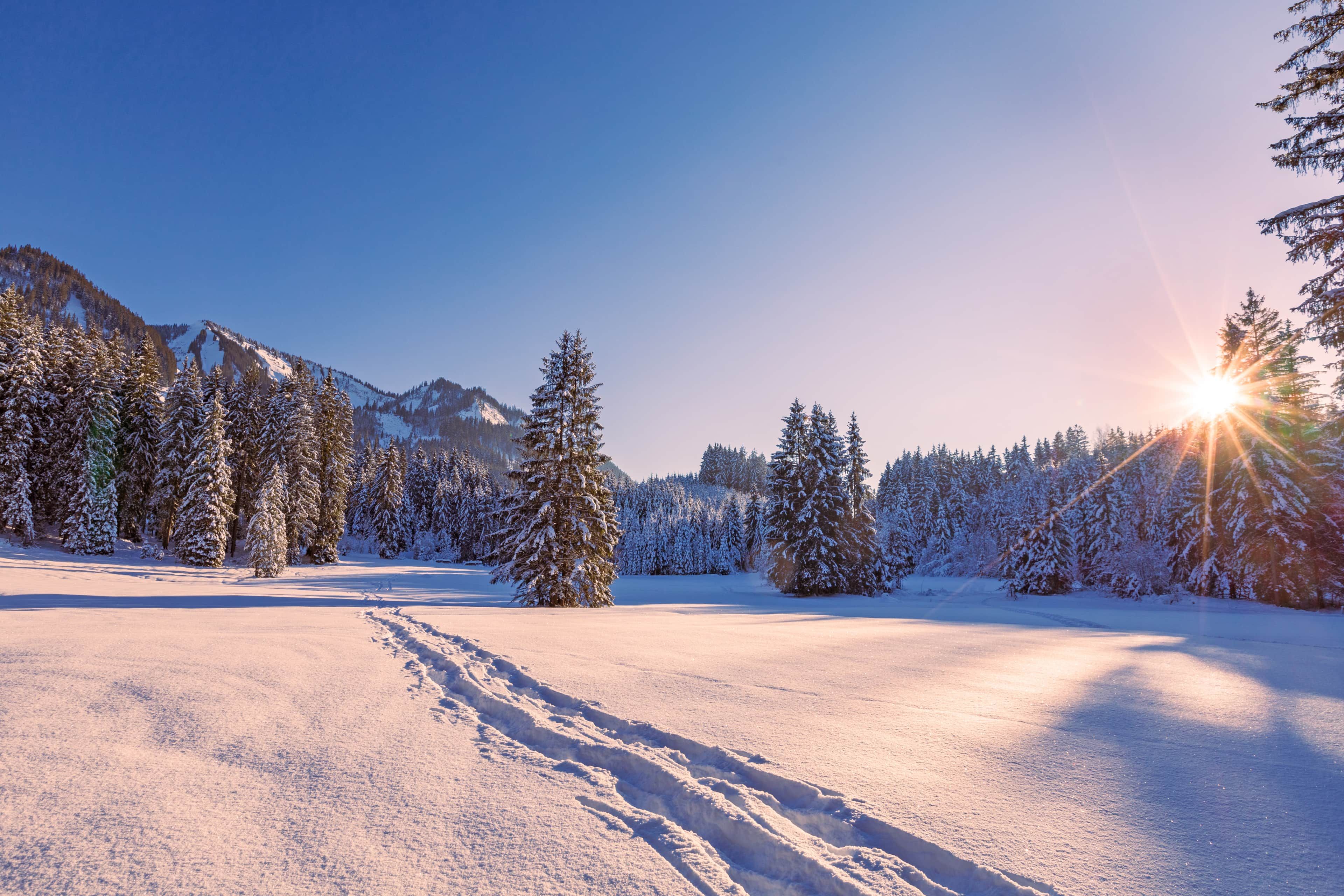 A snowy landscape with a clear sky and a sun setting or rising behind a mountain range.