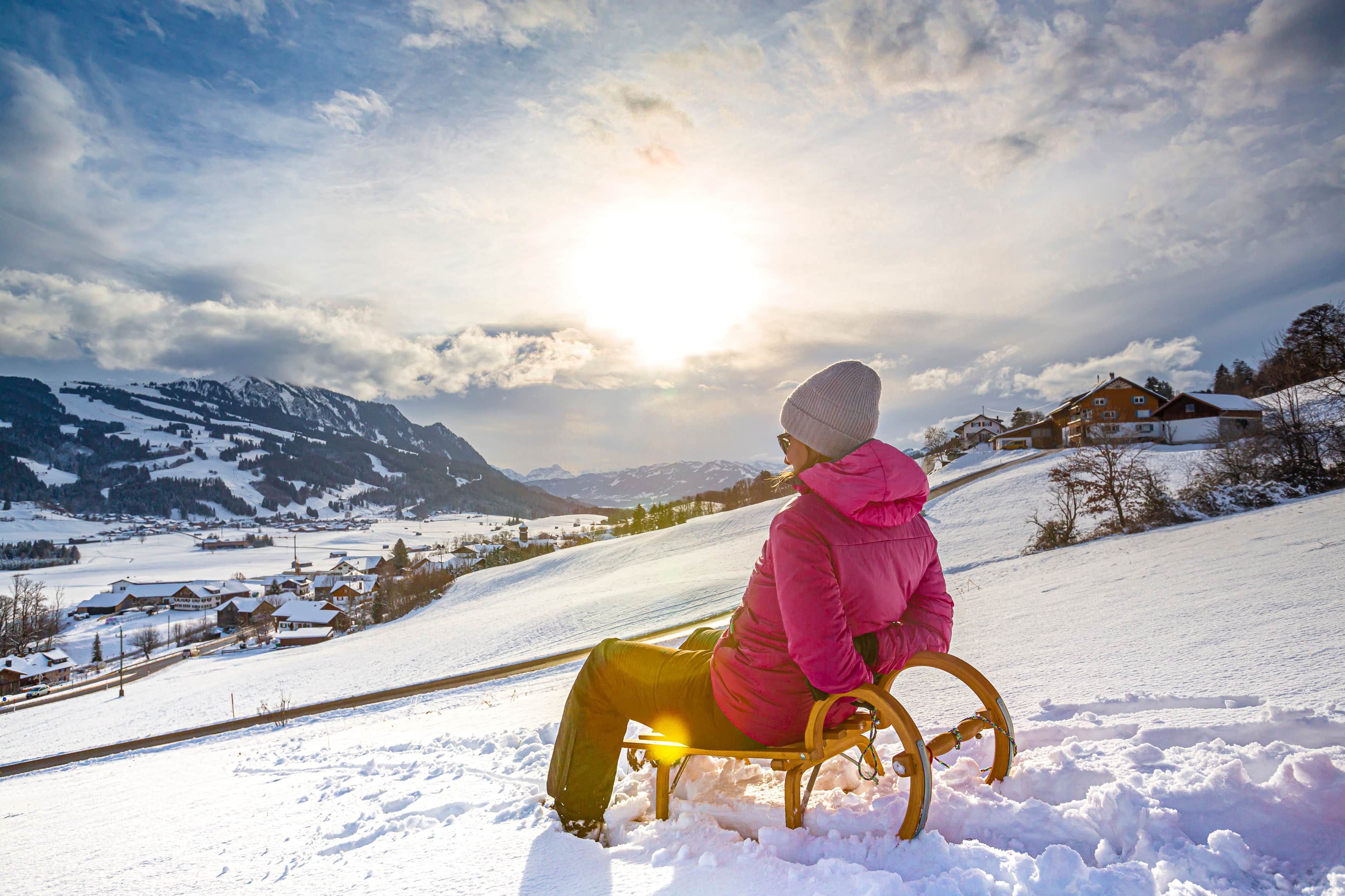 A person sitting on a wooden sled in a snowy landscape with a village and mountains in the background.