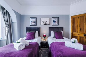 A hotel room with two beds, purple bedding, a nightstand with a lamp, and artwork on the wall. - NEW! Spacious Home, Ideal for Groups, FREE Parking (Manchester)