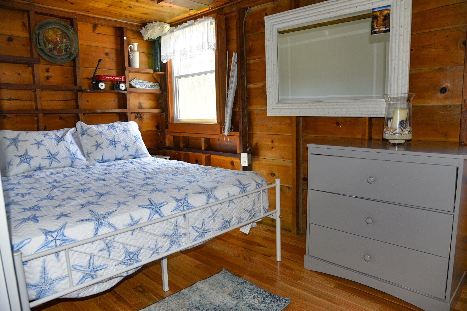 Une pièce chaleureuse avec un lit agrémenté d’une couvre-lit à motifs d’étoiles, une commode, une fenêtre avec des rideaux et un mur en bois orné d’une assiette décorative.