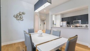 Un espace repas moderne avec une table blanche et des chaises grises, une cuisine dotée de meubles sombres et d’un four à micro-ondes, ainsi qu’un élément décoratif mural.