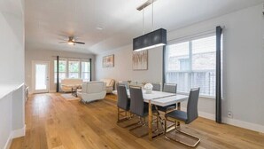 Un espace repas moderne avec une table et des chaises blanches, un ventilateur de plafond et une grande fenêtre équipée de stores.