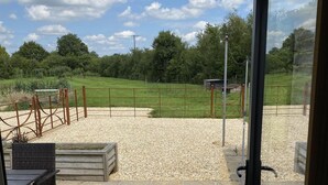 Blick aus einem Gebäude auf einen Kiesplatz mit einem Metallzaun, einer Holzbank und einem kleinen Schuppen im Hintergrund.