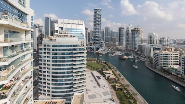 A high-rise hotel with multiple balconies, a marina with boats, and a city skyline in the background.