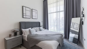 A modern bedroom with a large bed, a bedside table with a lamp, and a view of the outdoors through a window with curtains.