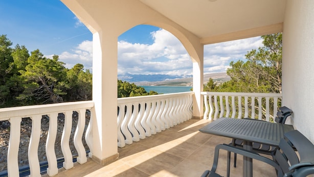 Una terraza techada con mesa y sillas, barandillas blancas y vistas al mar y las montañas.