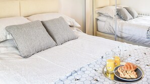 A neatly made bed with a patterned bedspread, pillows, a tray with a croissant and juice, and a table with a honeycomb tablecloth.
