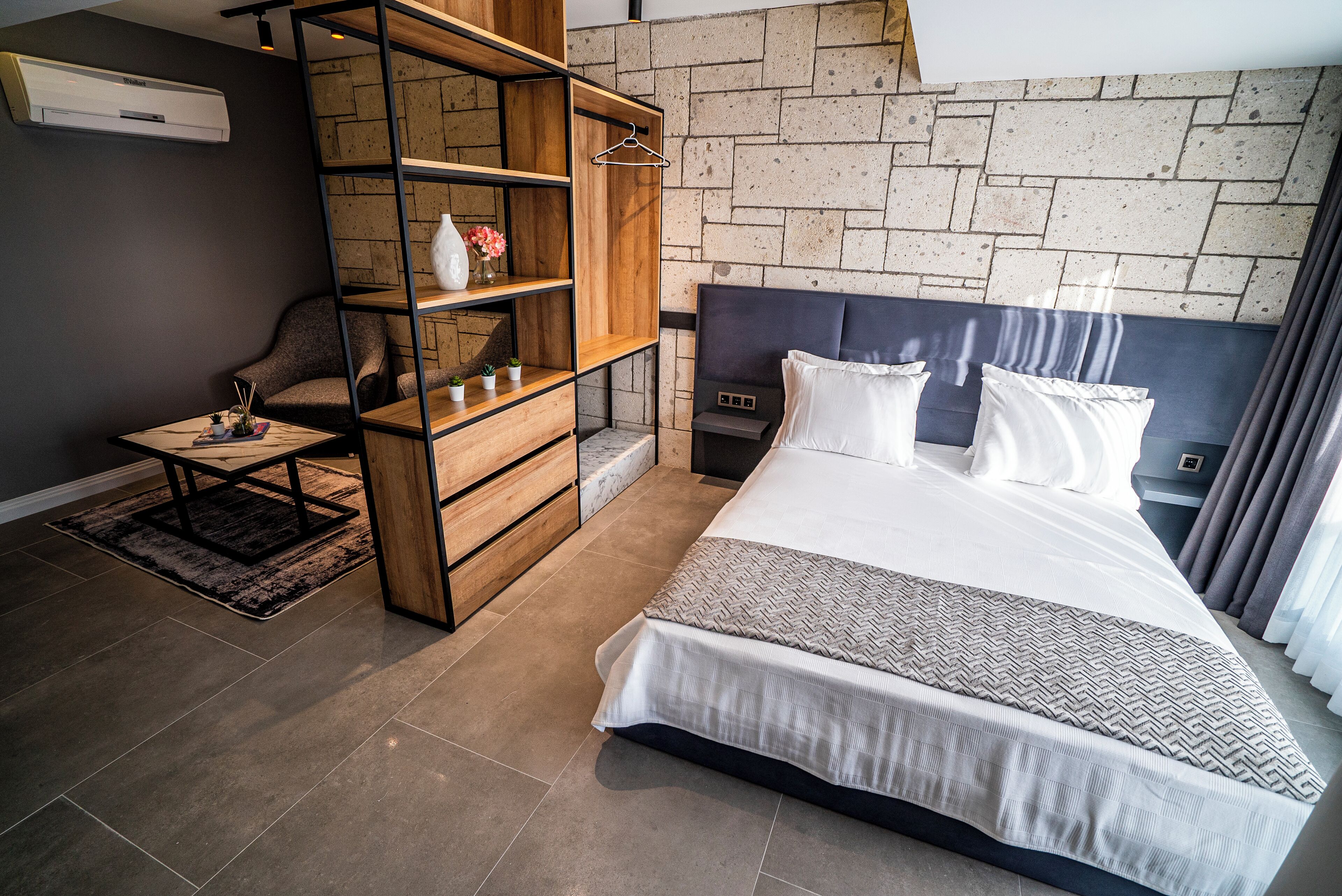 A modern bedroom with a large bed, a wooden shelving unit, a small table, and a brick wall.