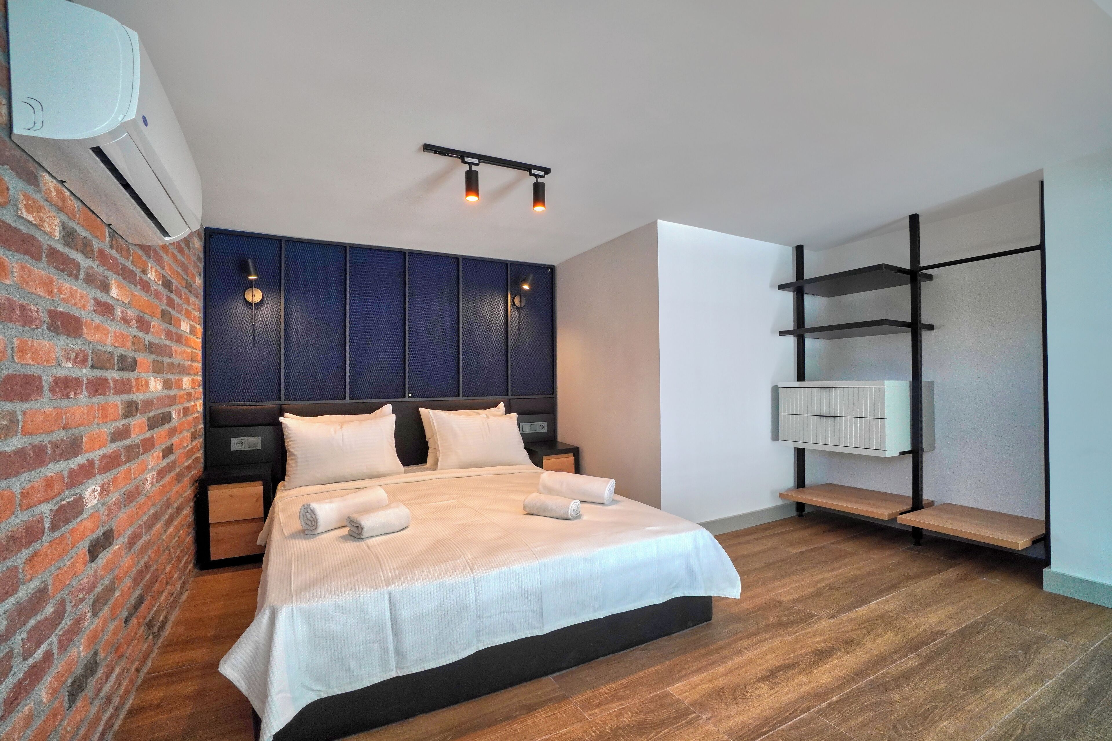 Une chambre moderne avec un mur en briques, un grand lit avec du linge de lit blanc, une tête de lit sombre et une armoire intégrée avec des étagères.