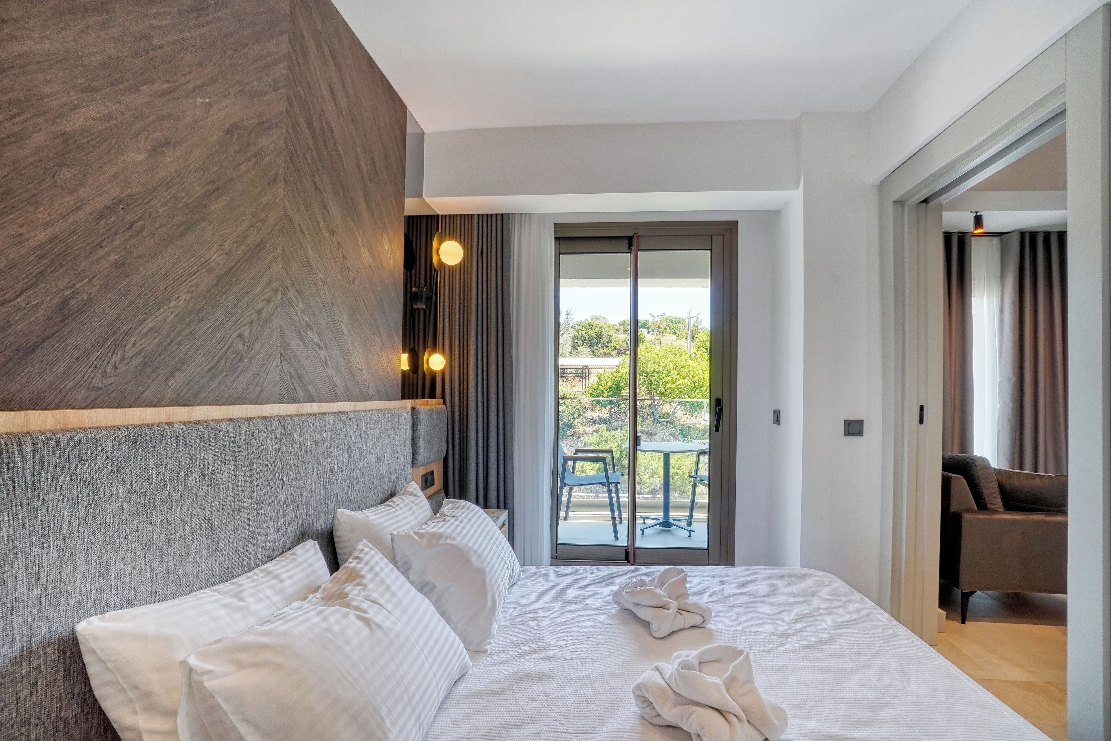 Habitación de hotel moderna con una cama grande, una pared decorativa de madera y un gran ventanal corredero que da a un balcón con mobiliario exterior.