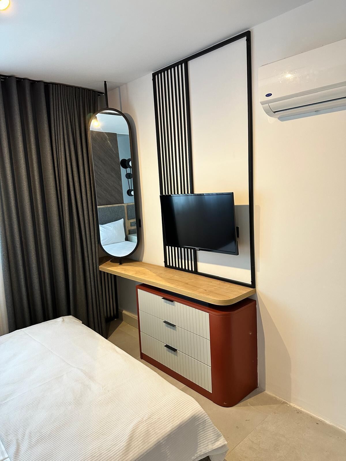Une chambre d’hôtel comprenant un lit, une table de chevet en bois avec des tiroirs, un grand miroir, un téléviseur à écran plat fixé au mur et un appareil de climatisation.