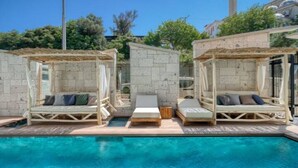 A poolside area with two wooden loungers under pergolas, a clear blue pool, and a mosque in the background. - Ayayorgi Homes & Suites (Cesme)
