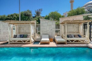 A poolside area with two wooden loungers under pergolas, a clear blue pool, and a mosque in the background.