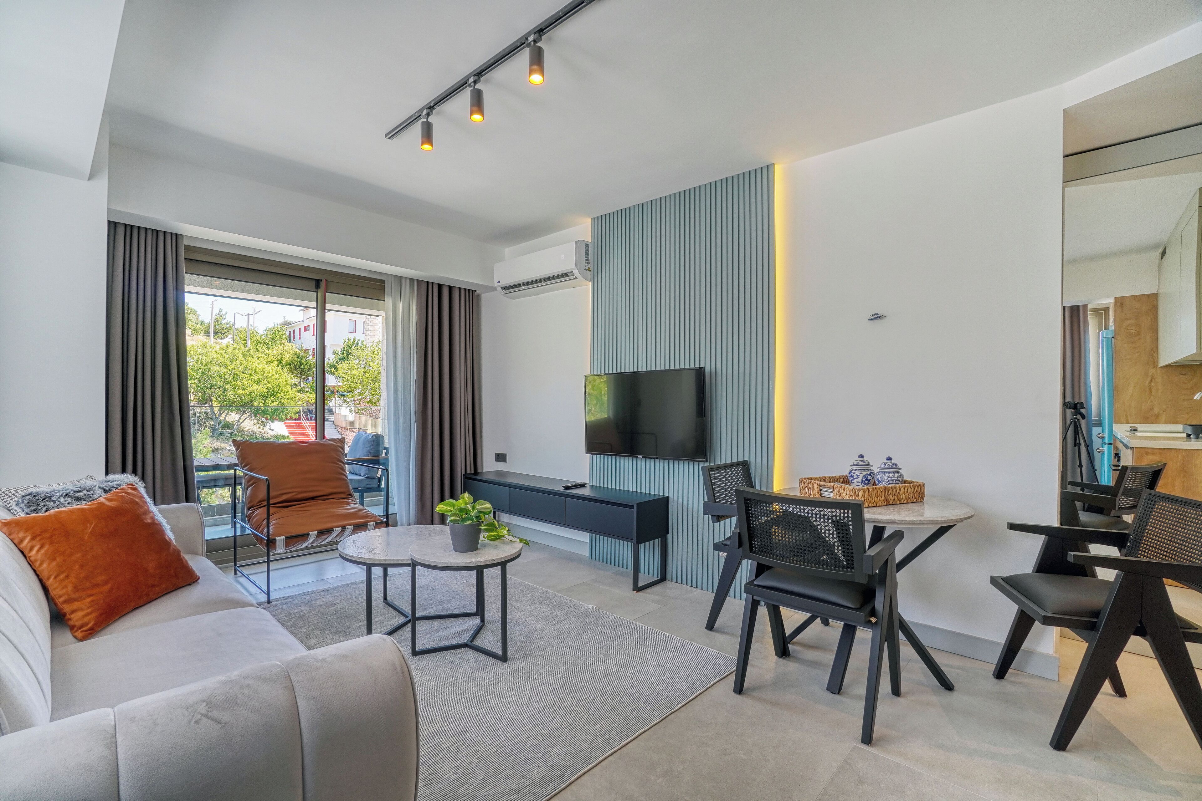 A modern living room with a sofa, a coffee table, and a TV. There is a balcony with curtains and a view of trees.