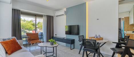 A modern living room with a sofa, a coffee table, and a TV. There is a balcony with curtains and a view of trees.