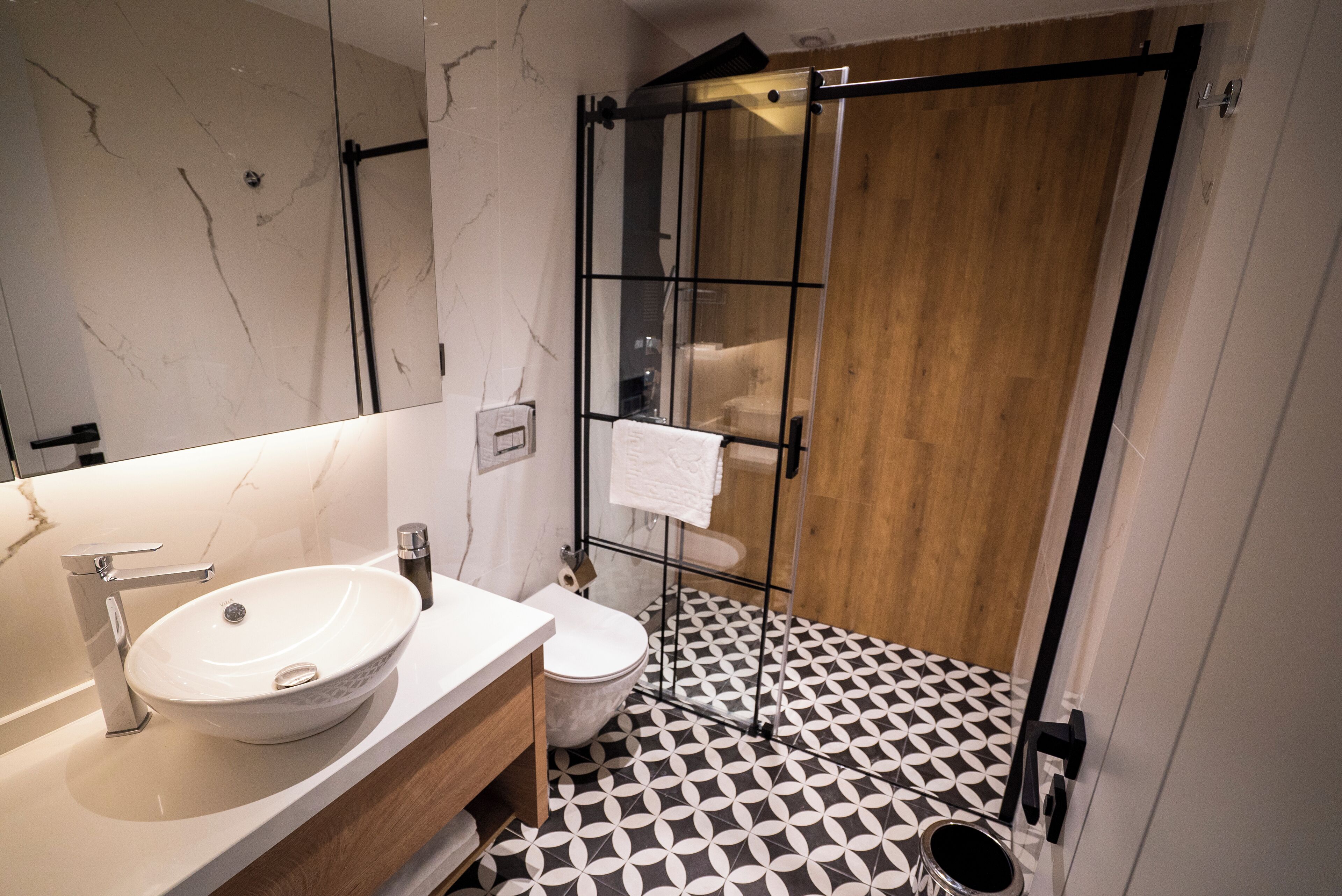 Un baño moderno con lavabo de mármol blanco, inodoro y ducha con cerramiento de vidrio. El piso tiene un estampado geométrico blanco y negro.