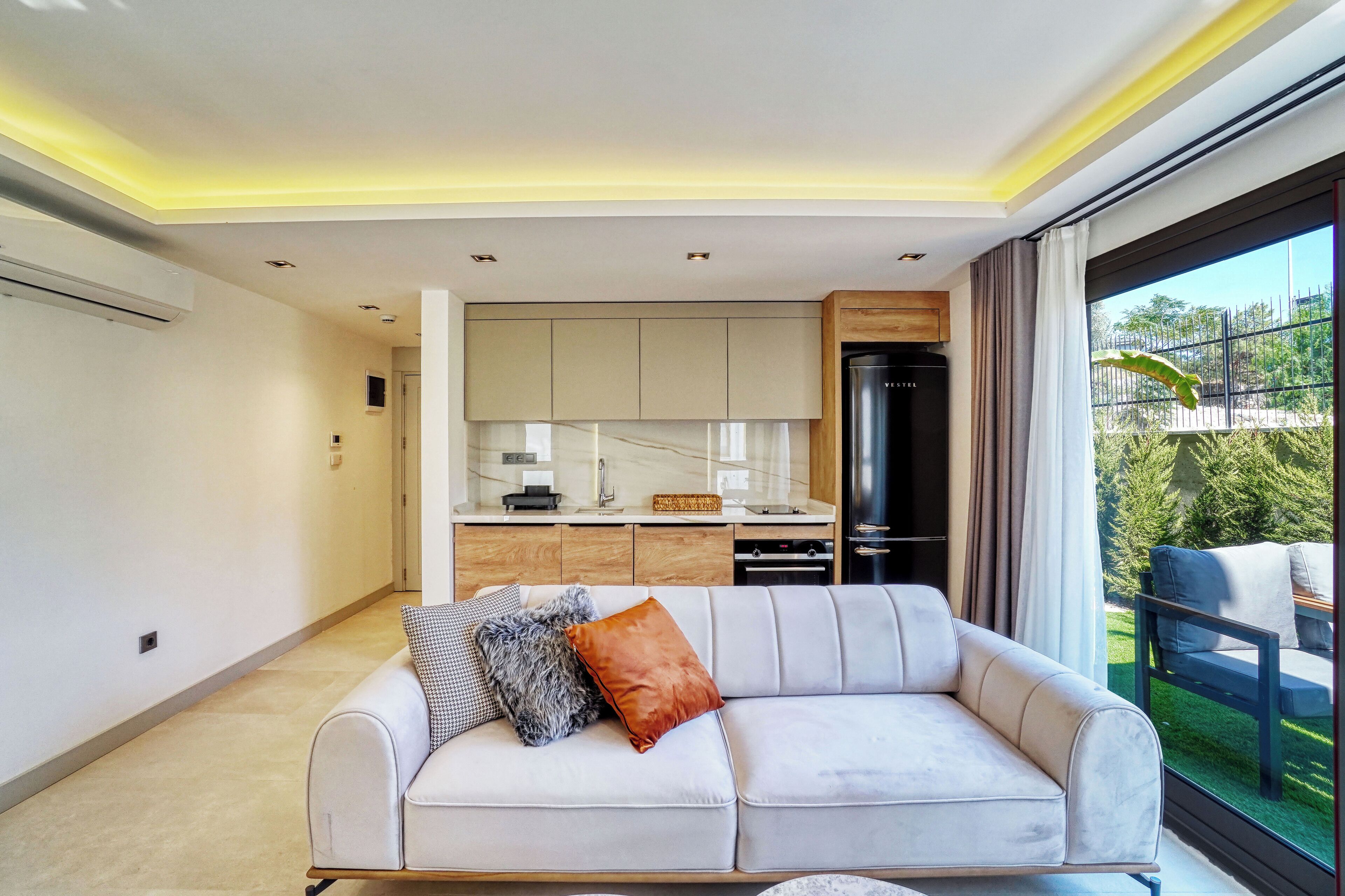 A modern living room with a sofa, a kitchen area, and a large window with a view of greenery.