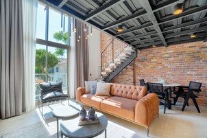 A modern living room with a leather sofa, a round coffee table, and a dining area with chairs and a table. A brick wall and a staircase are visible in the background.