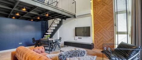 A modern living room with a sofa, ottoman, and a black chair. There is a staircase leading to an upper level, a dining area, and a TV mounted on the wall.