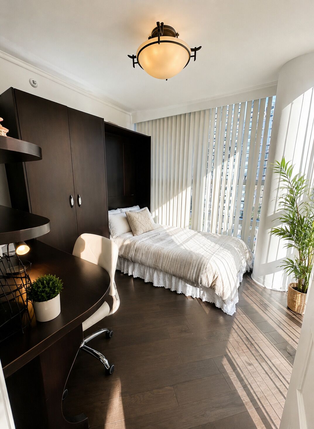 A bedroom with a bed, a desk, a chair, a potted plant, and a window with blinds.
