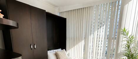 A bedroom with a bed, a desk, a chair, a potted plant, and a window with blinds.