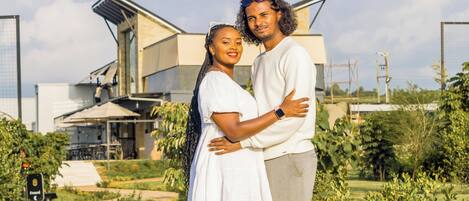 Una pareja abrazándose en un entorno exterior moderno con una casa y vegetación al fondo.