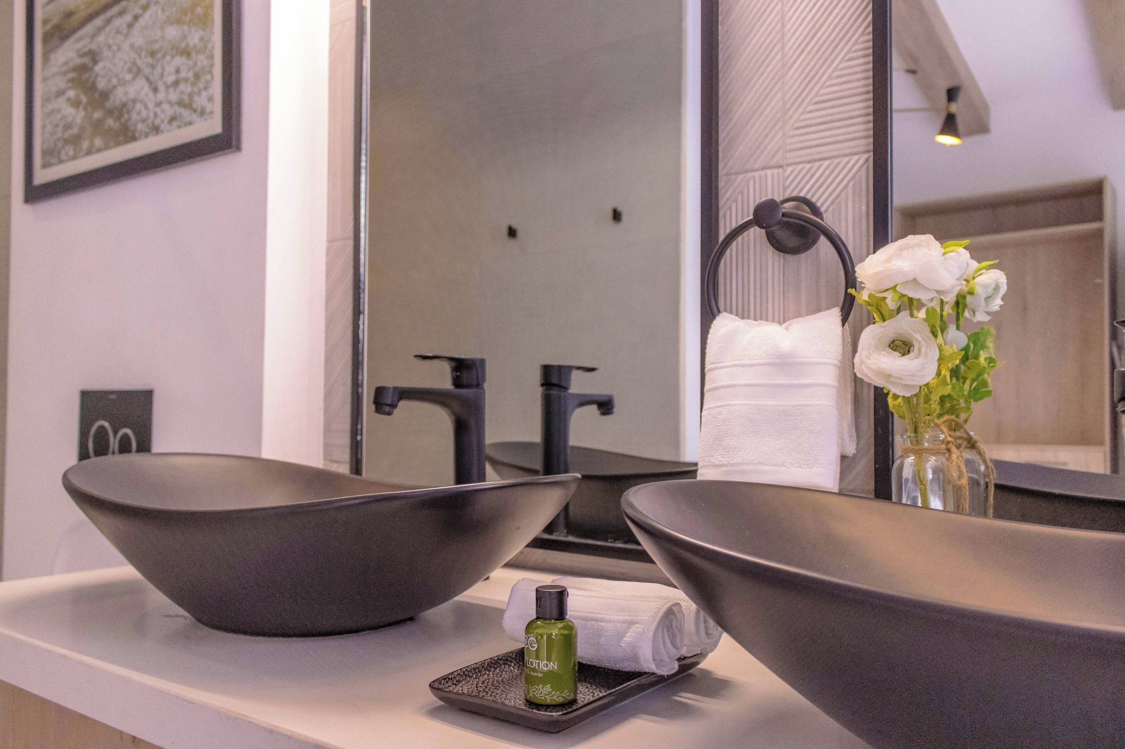 A bathroom with two large, dark bowls as sinks, a small bottle of lotion, and a vase with white flowers.