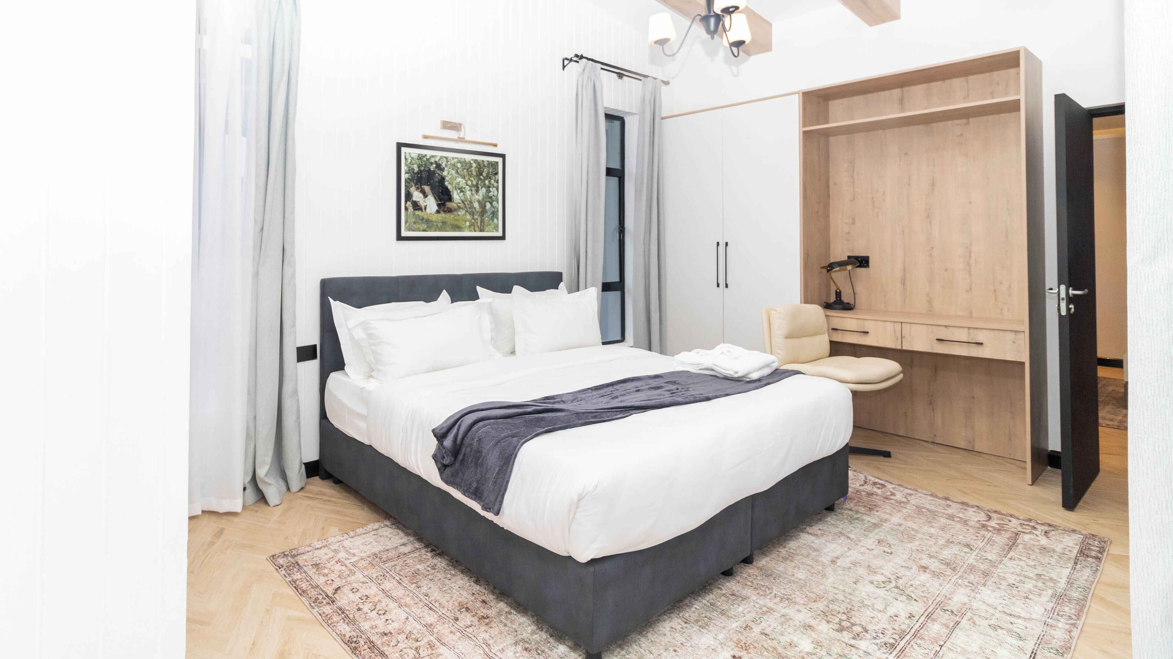 A modern bedroom with a large bed, a wooden wardrobe, a small desk, and a framed picture on the wall.