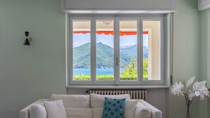 Un sofá blanco con un cojín estampado, una ventana con vistas a montañas y vegetación, y un florero con flores blancas.